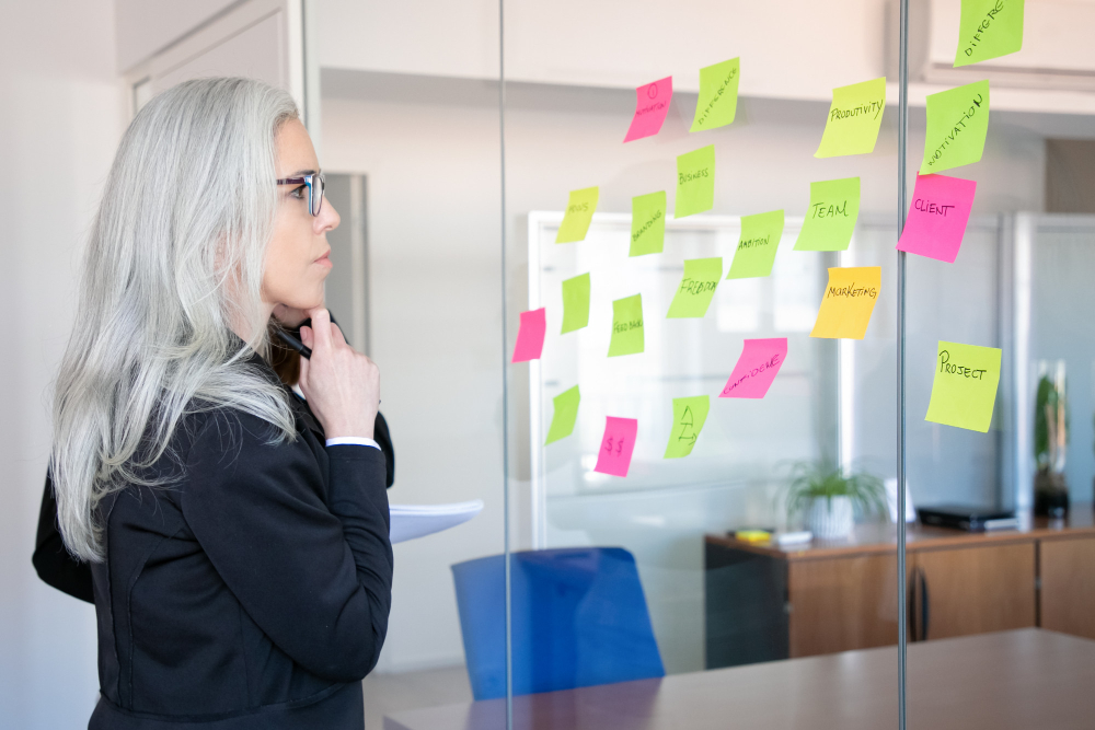confident-concentrated-businesswoman-looking-at-stickers-on-glass-wall-focused-grey-haired-female-worker-thinking-about-notes-for-project-strategy-marketing-business-and-management-concept